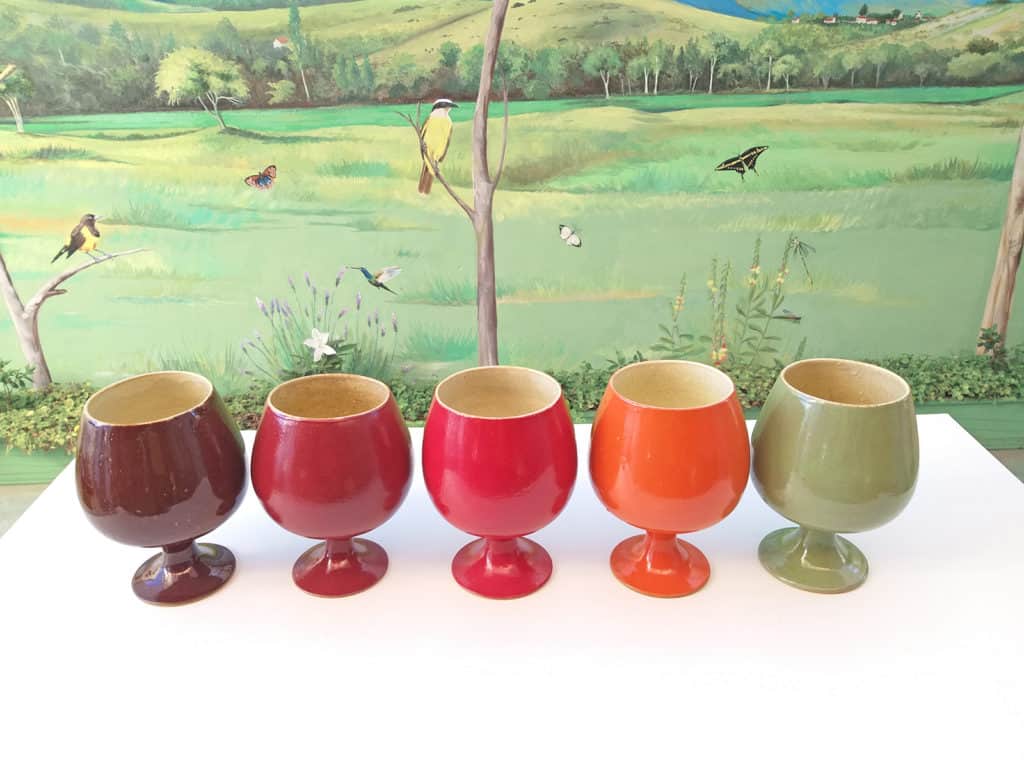 Cinco taças de cerâmica em cores diferentes: marrom, vinho, vermelho, laranja e verde. Ao fundo, vemos uma pintura mural de paisagem verdejante com pássaros