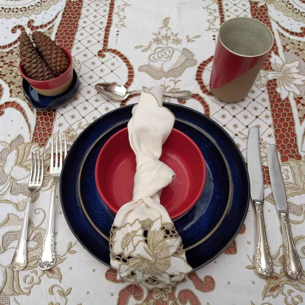 Foto feita de cima mostra lugar na mesa posta de Natal com pratos em tom de azul ou vermelho. A toalha é de linho bordado, bege.