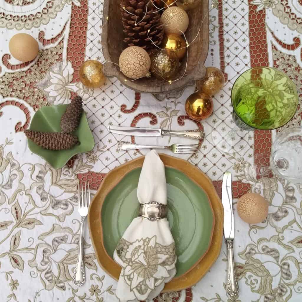 Foto feita de cima mostra lugar na mesa de Natal com pratos de formas orgânicas em tom de verde e bege. A toalha é de linho bordado, bege.