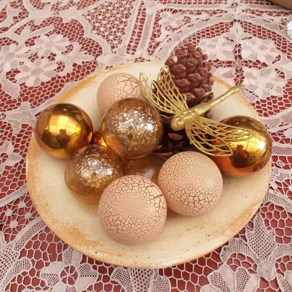 Prato fundo bege com bolas de Natal douradas, uma pinha e uma libélula dourada de plástico. 