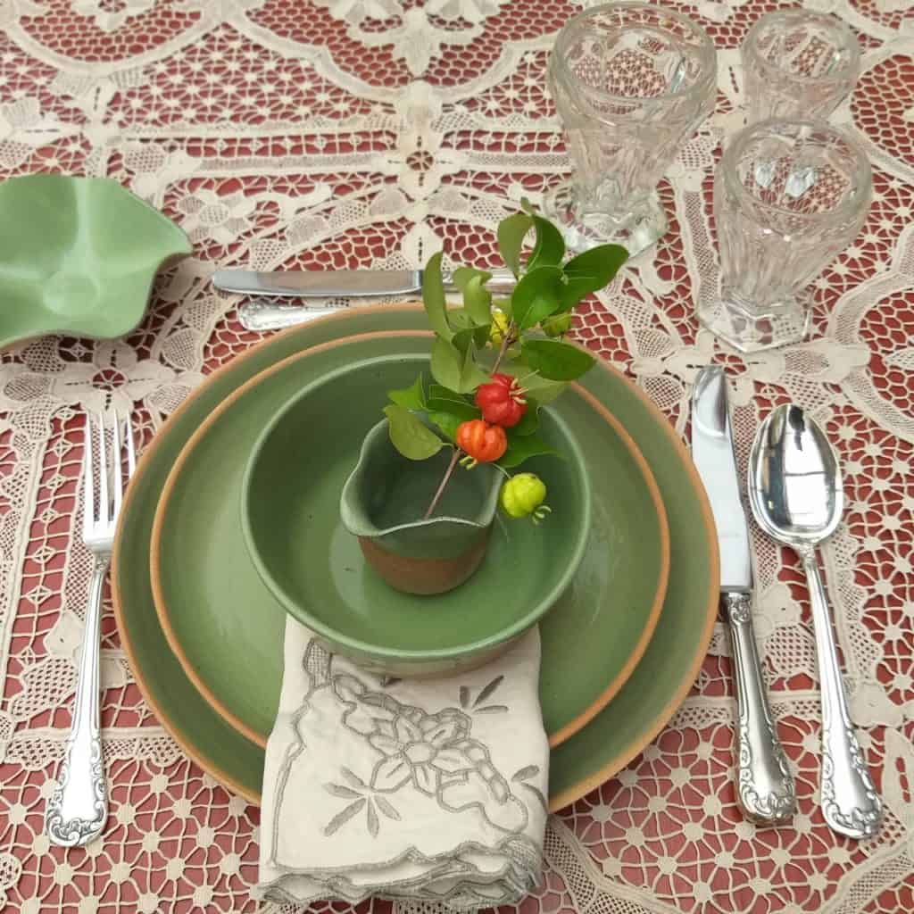 Foto feita de cima mostra lugar na mesa posta de Natal com pratos em tom de verde. A toalha é de renda artesanal, bege.