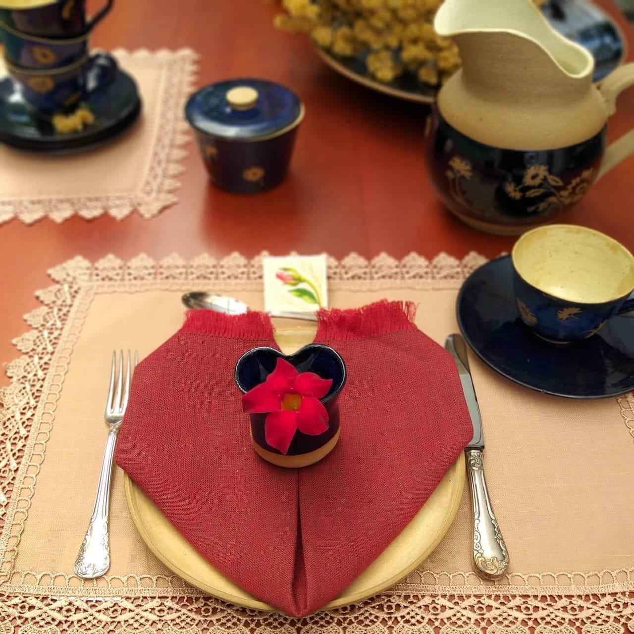 Lugar na mesa posta para o Dia das Mães com prato de cerâmica bege, xícara, jarra e manteigueira em bege e azul escuro, com desenhos de flores. Há um guardanapo vermelho, dobrado em forma de coração, em cima do prato. 