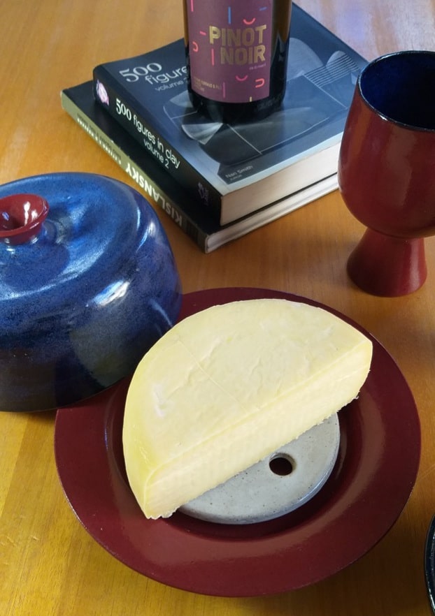 Queijeira de cerâmica com tampa azul apoiada sobre prato vermelho escuro onde temos metade de um queijo curado. Ao fundo vemos uma garrafa de vinho tinto. E à direita, uma taça de cerâmica vermelha. 
