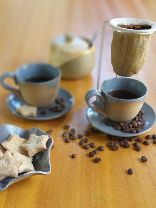 Duas xícaras de cafezinho, de cerâmica bege e cinza. Há um mini coador de pano sobre uma delas. Ao fundo, vemos um açucareiro . E à frente, do lado esquerdo, há um pratinho com biscoitos. Vários grãos de café estão espalhados sobre a mesa.