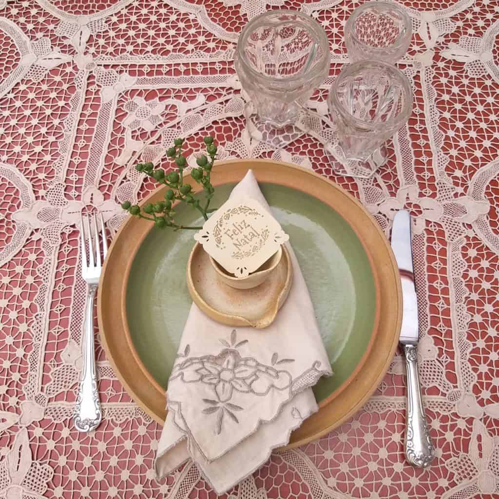 Lugar na mesa posta com prato de sobremesa verde claro sobre prato raso marfim, ambos de cerâmica. a toalha de renda e o guardanapo de linho são beges. 