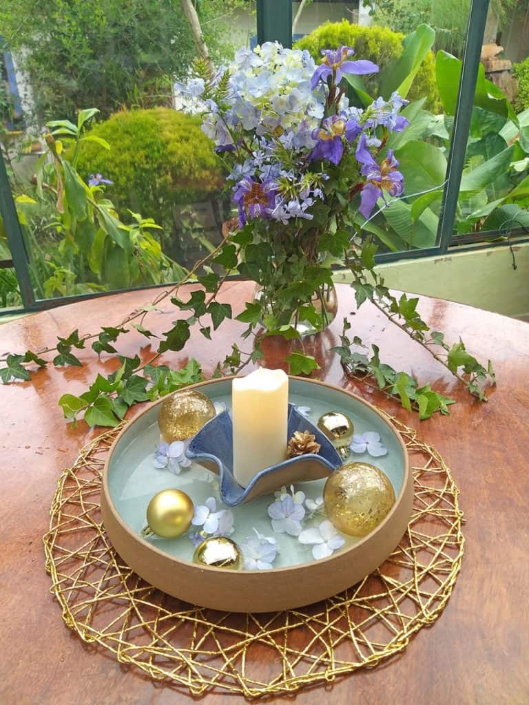 No centro da mesa, há uma travessa redonda de cerâmica, com o interior em azul claro. Dentro dela há água, bolas de Natal douradas, flores azuis, e um pratinho azul contendo uma vela. 